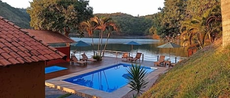 A pool area with lounge chairs and umbrellas, surrounded by lush greenery and a view of a lake.