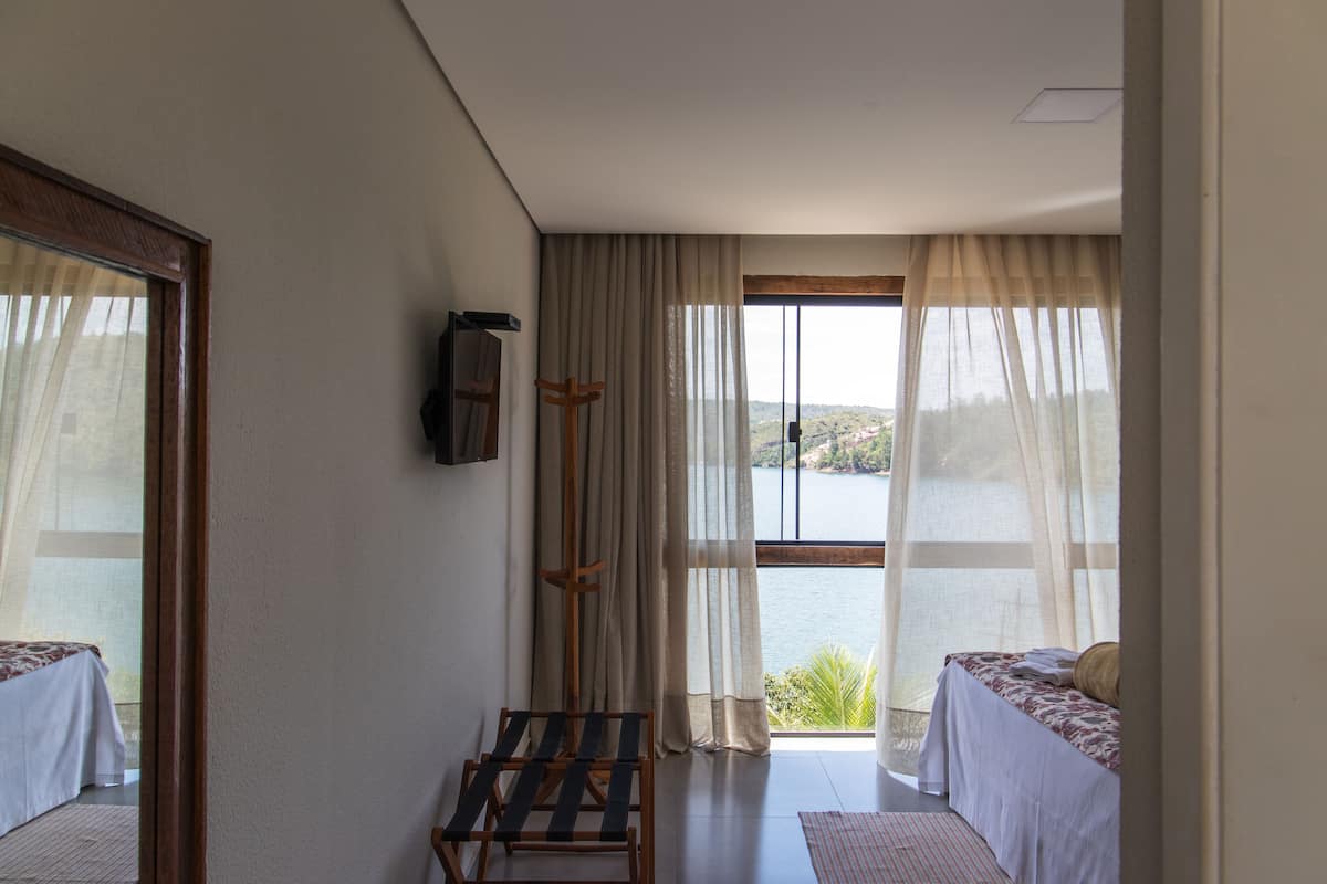 Quarto com duas camas, uma cadeira de madeira, janela com cortinas leves e vista para um corpo d'água.