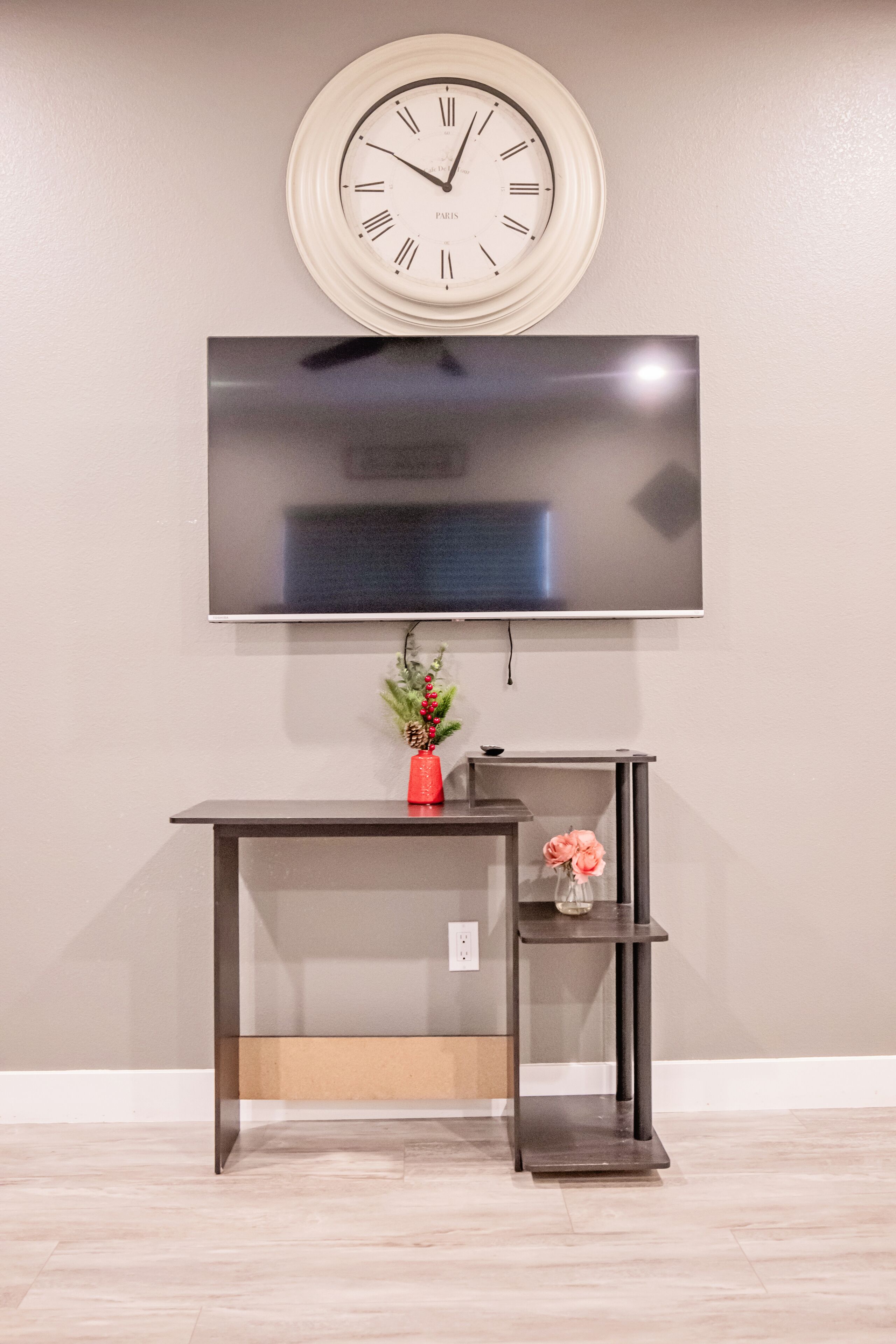 En moderne stue med et vægmonteret fladskærms-tv, et vægur og et lille skrivebord med en vase blomster.