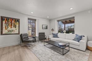 Una sala de estar moderna con un sofá blanco, sillones de cuero, una mesa de centro de vidrio y un ventanal con vistas a árboles.