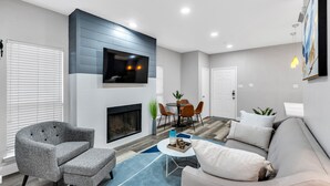 Un salon moderne avec une cheminée, des meubles gris et blancs, un téléviseur à écran plat fixé au mur et un coin repas avec une table et des chaises.