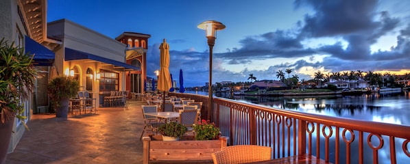 An outdoor patio with tables and chairs, a view of a body of water, and a building with a balcony.