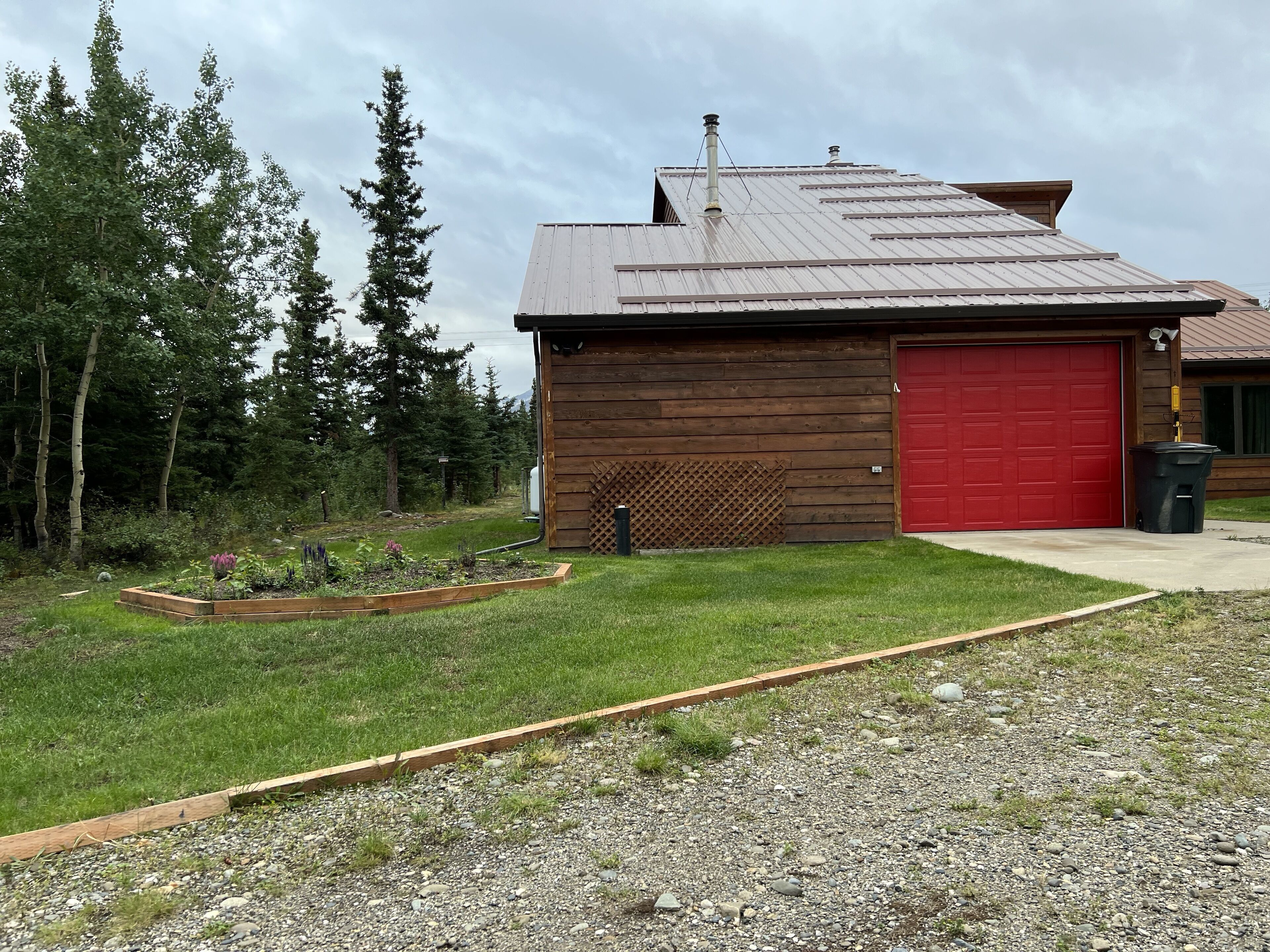 A wooden cabin with a red garage door, surrounded by a garden and trees.