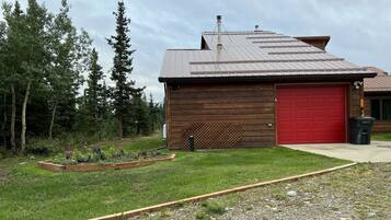 A wooden cabin with a red garage door, surrounded by a garden and trees.
