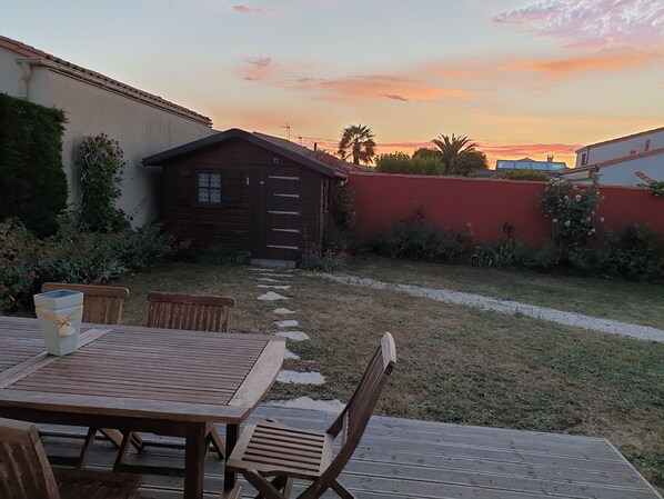 A wooden deck with a table and chairs, a small wooden cabin, and a red wall with greenery.