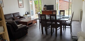 A living room with a sofa, a coffee table, and a dining table with chairs. There is a television on a wooden cabinet and a window with a view of a garden.
