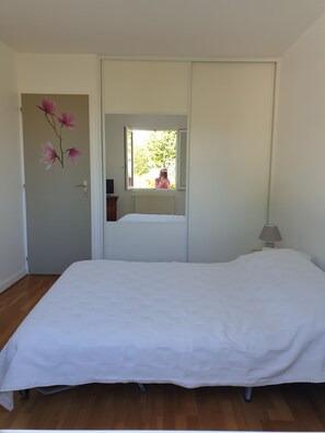 A bedroom with a large bed, a wardrobe, a window with a view of greenery, and a small table with a lamp.