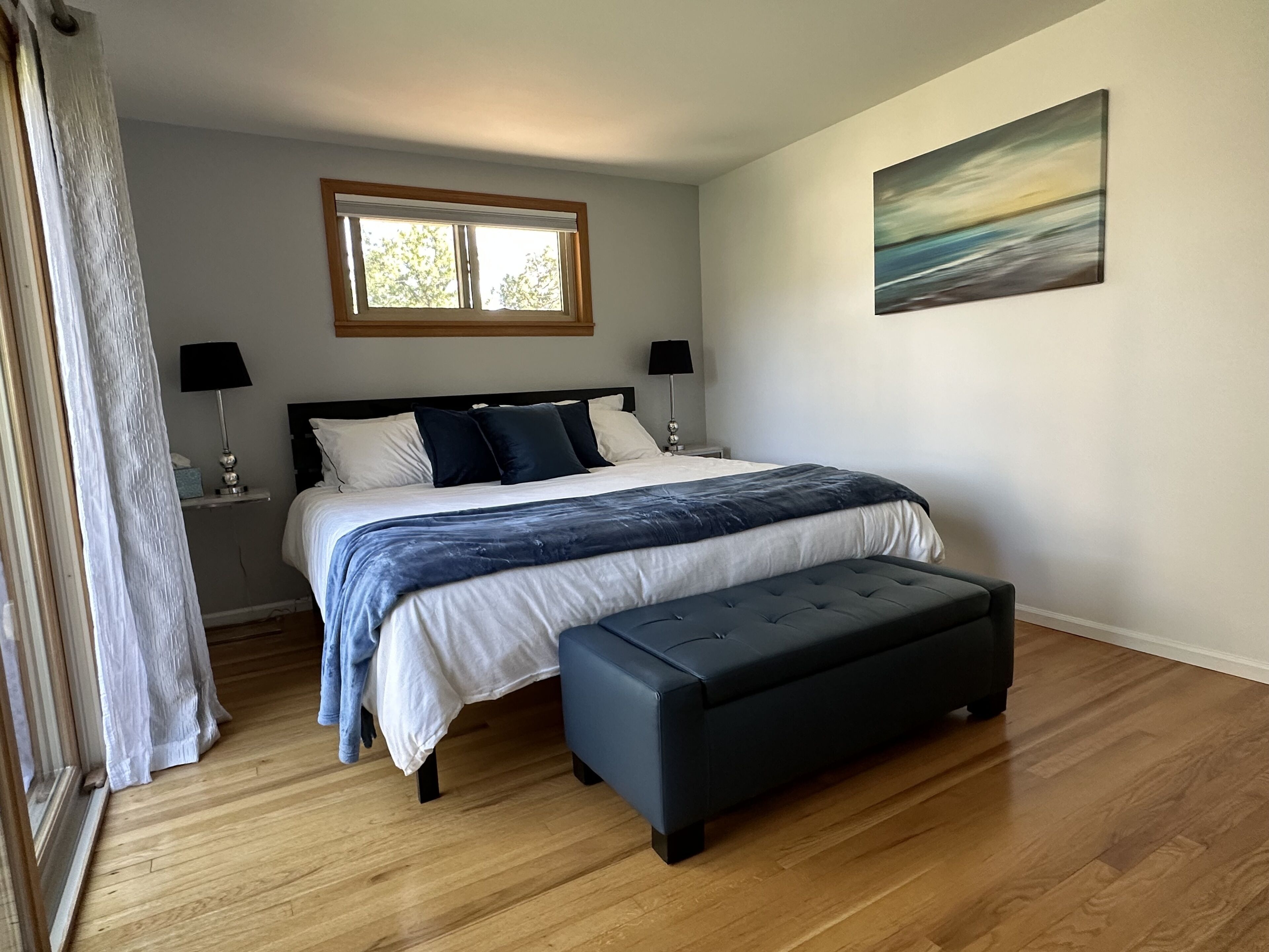 A bedroom with a bed, bedside lamps, a window, a painting, and a bench.