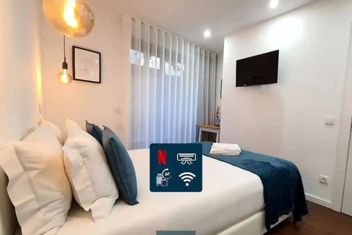 A modern hotel room with a neatly made bed, a flat-screen TV mounted on the wall, and a large window with vertical blinds.