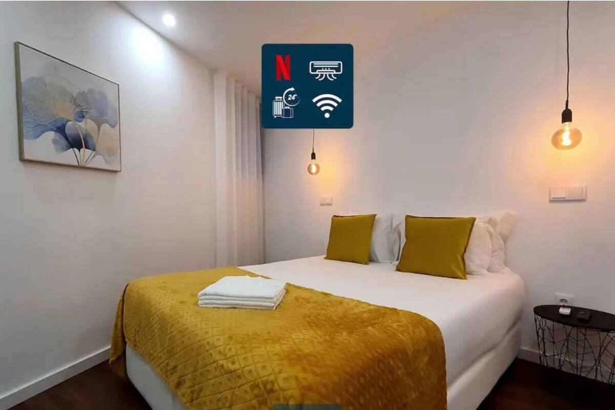 A hotel room with a bed, a yellow throw blanket, a painting on the wall, and a sign indicating amenities like Netflix, laundry, and Wi-Fi.