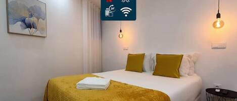 A hotel room with a bed, a yellow throw blanket, a painting on the wall, and a sign indicating amenities like Netflix, laundry, and Wi-Fi.
