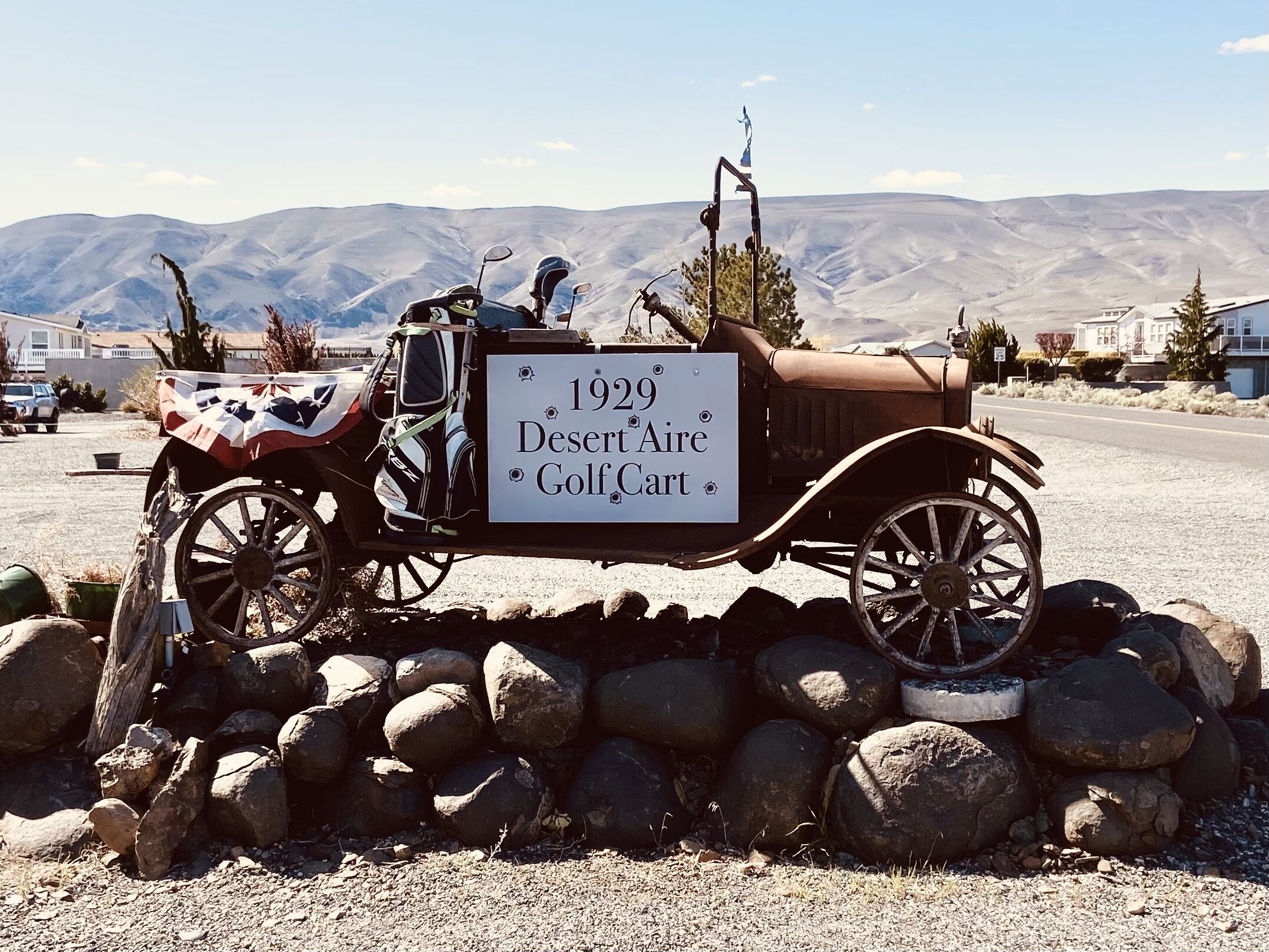 A vintage 1929 Desert Aire Golf Cart parked on a gravel surface with a signboard, surrounded by rocks and a mountainous backdrop.