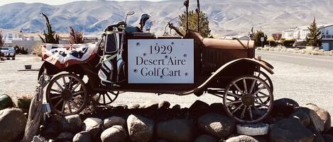 A vintage 1929 Desert Aire Golf Cart parked on a gravel surface with a signboard, surrounded by rocks and a mountainous backdrop.
