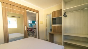 A bedroom with a bed, a wooden wardrobe, and a kitchenette in the background.