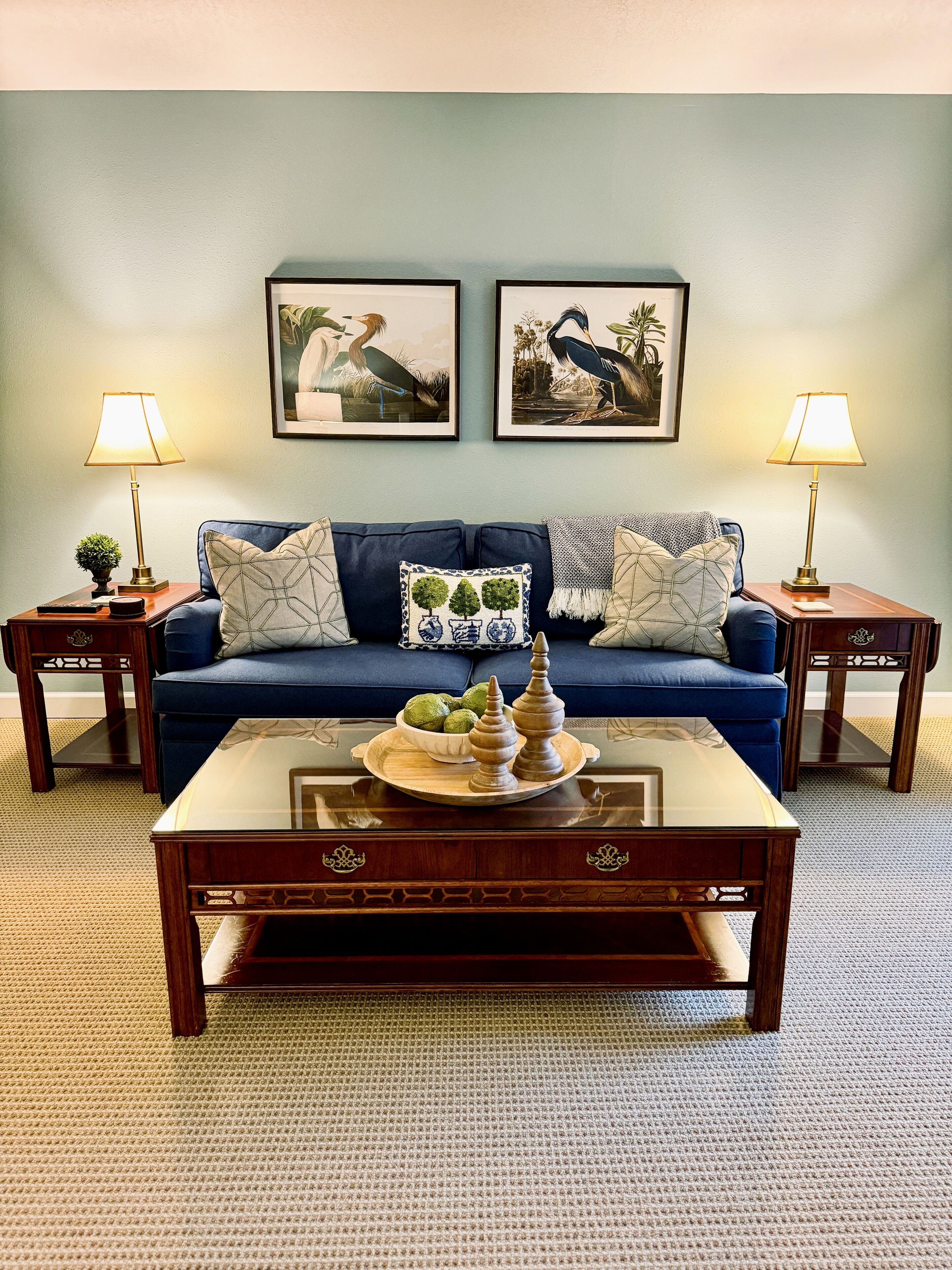 A living room with a blue sofa, two side tables with lamps, a coffee table with decorative items, and two framed wall art pieces.