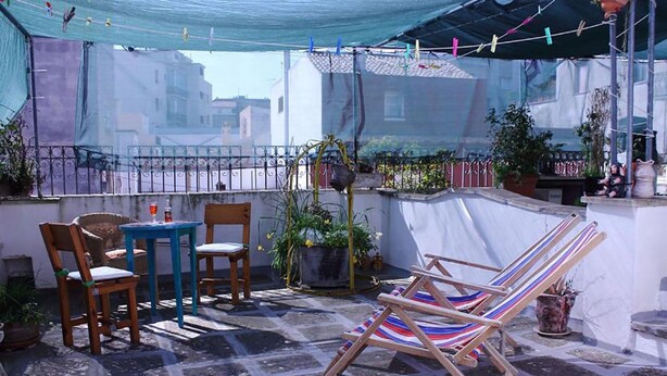 Een terras met een tafel en stoelen, een gestreepte strandstoel, planten in potten en een uitzicht op gebouwen.
