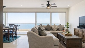 A modern living room with a large window, a ceiling fan, a sofa, a coffee table, and a TV. - 6th-Floor Oceanfront Condo - Indoor Pools, Sauna, Private Beach (Rosarito BC)
