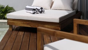 A sun lounger with a white cushion and a black and white pillow, positioned on a wooden deck with a view of a building and a plant in the background. - Neptune Studios Lombok (Kuta)