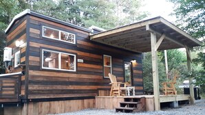 A cabin with a covered porch, wooden exterior, and two windows. - Big Porch Tiny House (Brevard)