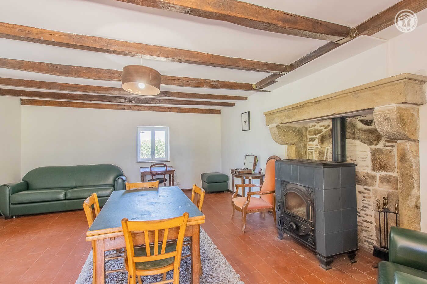 A cozy room with a stone fireplace, wooden beams, a dining table with chairs, a green sofa, and a window.