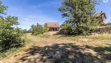 Ein rustikales Haus mit Steinmauer und Holzdach, umgeben von Bäumen und einem trockenen, grasbewachsenen Feld.