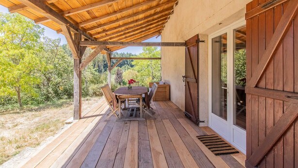 A covered outdoor area with a wooden table and chairs, a view of the surrounding trees, and a door leading to the interior. - Thélème - Pouilloux - Gîte (POUILLOUX)