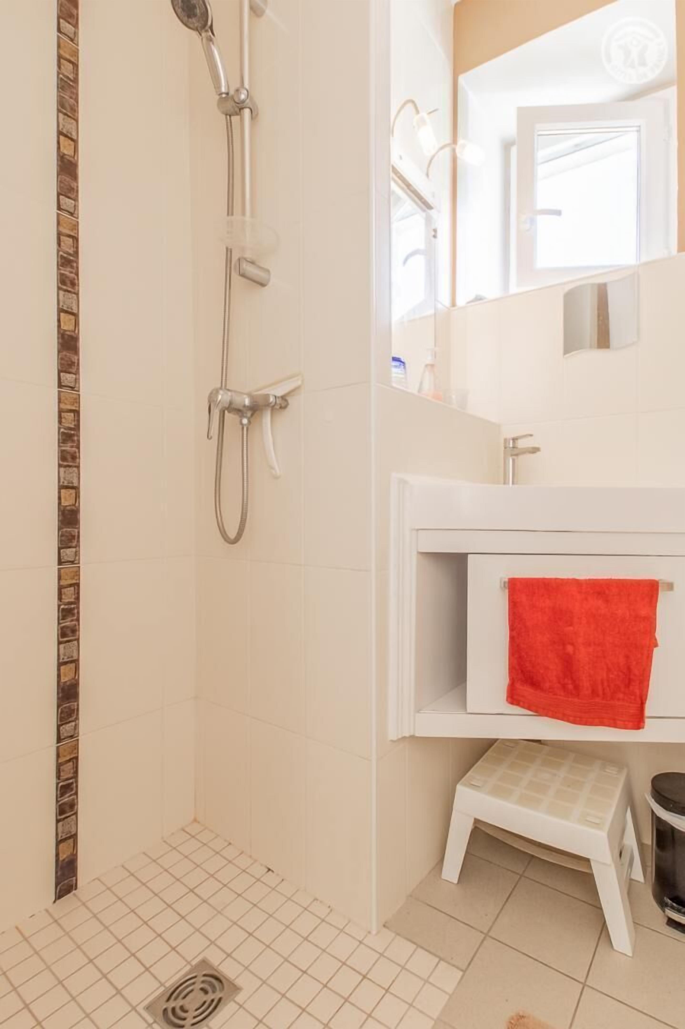 A bathroom with a shower, a white sink with a red towel, and a small step stool.