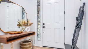 A hallway with a white door, a wooden console table with a mirror, a wicker basket, and a black umbrella leaning against the wall. - New 4br home 5 min from the beach-Paradise escape (Waianae)