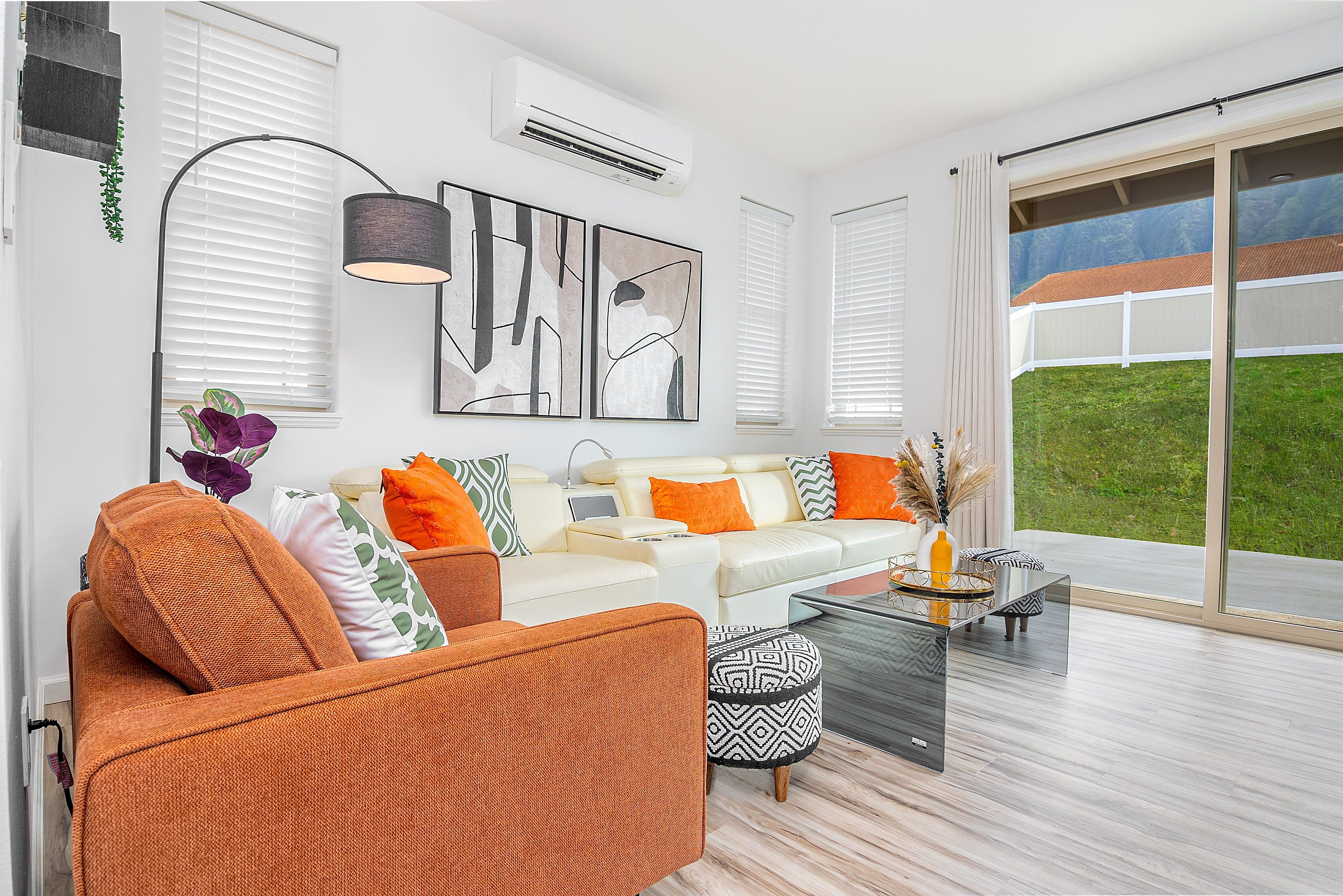 A modern living room with a white sofa, orange armchair, and patterned ottoman. There is a glass coffee table, a floor lamp, and artwork on the wall.
