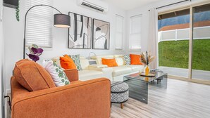 A modern living room with a white sofa, orange armchair, and patterned ottoman. There is a glass coffee table, a floor lamp, and artwork on the wall. - New 4br home 5 min from the beach-Paradise escape (Waianae)