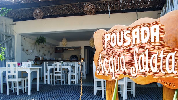 Ein Essbereich im Freien mit weißen Tischen und Stühlen, einem Schild mit der Aufschrift 'Pousada Acqua Salata' und einem strohgedeckten Bauwerk.