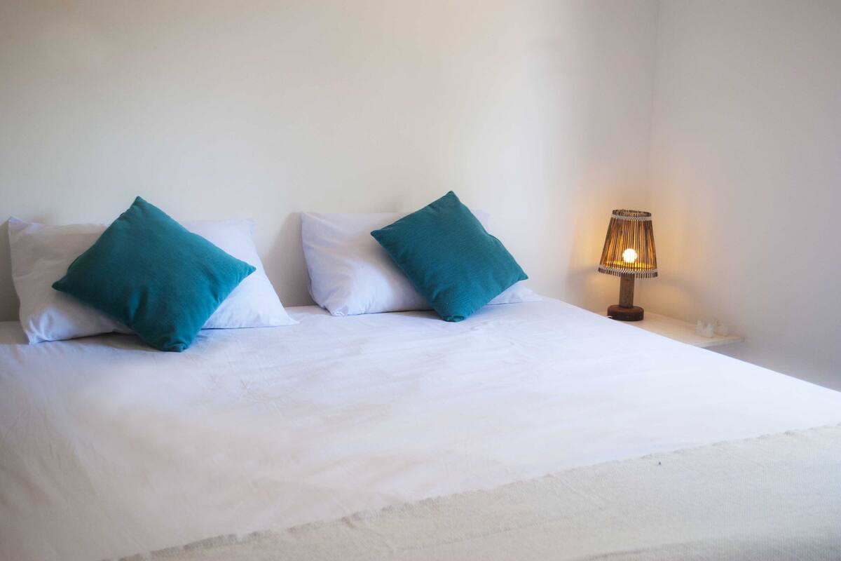 Una cama bien hecha con ropa de cama blanca y dos almohadas color turquesa, una mesita de noche pequeña con una lámpara, y una pared blanca lisa.
