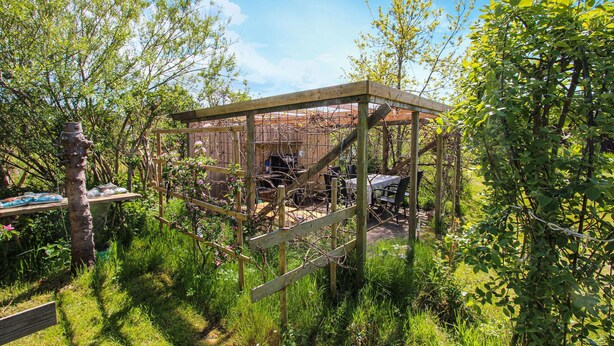 Een houten pergola in een tuin, met een tafel en stoelen eronder.