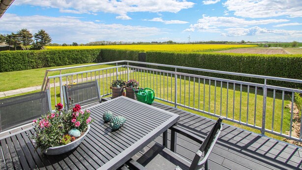 A balcony with a wooden table, potted plants, and a view of a field and hedge.
