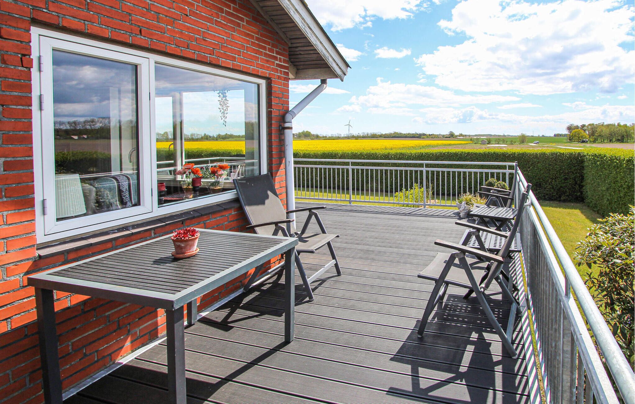 A deck with a table and chairs, a potted plant, and a view of a field and wind turbine.