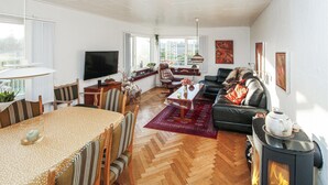 A spacious living room with a wooden herringbone floor, a dining area with a wooden table and chairs, a black leather sofa, a fireplace, and a large window with a view of the city.
