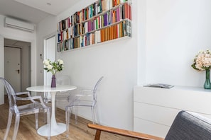 A room with a white wall, a bookshelf filled with books, a round white table with clear chairs, and a white cabinet.