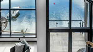 A modern interior with large windows overlooking a calm body of water, a small seating area with a cushion, and a metal railing on the deck.