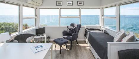 Une petite pièce comprenant un lit, un bureau et une chaise, le tout disposé face à une grande fenêtre donnant sur la mer.