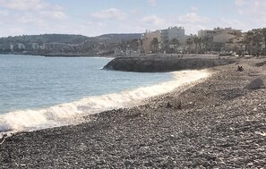Ein felsiger Strand mit brechenden Wellen, eine Steinmauer und Gebäude im Hintergrund.