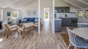 Amplia sala de estar con un sofá, sillas y una mesa de comedor. Se ve una cocina con armarios y electrodomésticos al fondo.