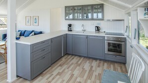 Una cocina moderna con armarios grises, encimera blanca y horno integrado. Se ve una ventana en el lado derecho, y en el fondo se distingue un comedor con mesa y sillas.