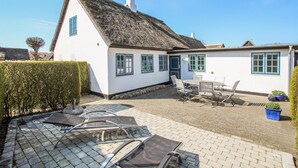 Une maison blanche avec un toit de chaume, une terrasse avec des chaises et une table en métal, et une haie au premier plan.