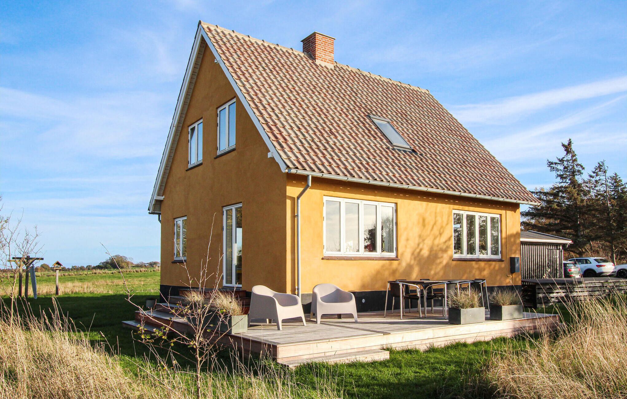 A yellow house with a tiled roof, a deck with chairs, and a car parked nearby.
