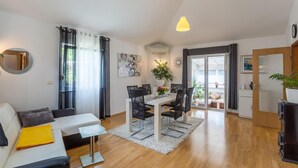 A modern living room with a sofa, a dining table with chairs, a small side table, a hanging light fixture, and a view through an open door to an outdoor area.