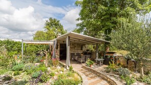 A covered outdoor dining area with stone walls, surrounded by a garden with various plants and a stone pathway.