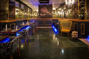 Uma área de bar com um balcão comprido, várias mesas e cadeiras. Há um espelho grande na parede e um lustre.