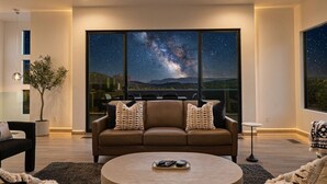 A modern living room with a large window displaying a night sky view, a brown leather sofa, a round coffee table, and a potted plant.