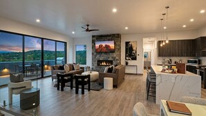 A modern living room with a fireplace, a kitchen with dark wood cabinets, and a large window overlooking a scenic landscape.