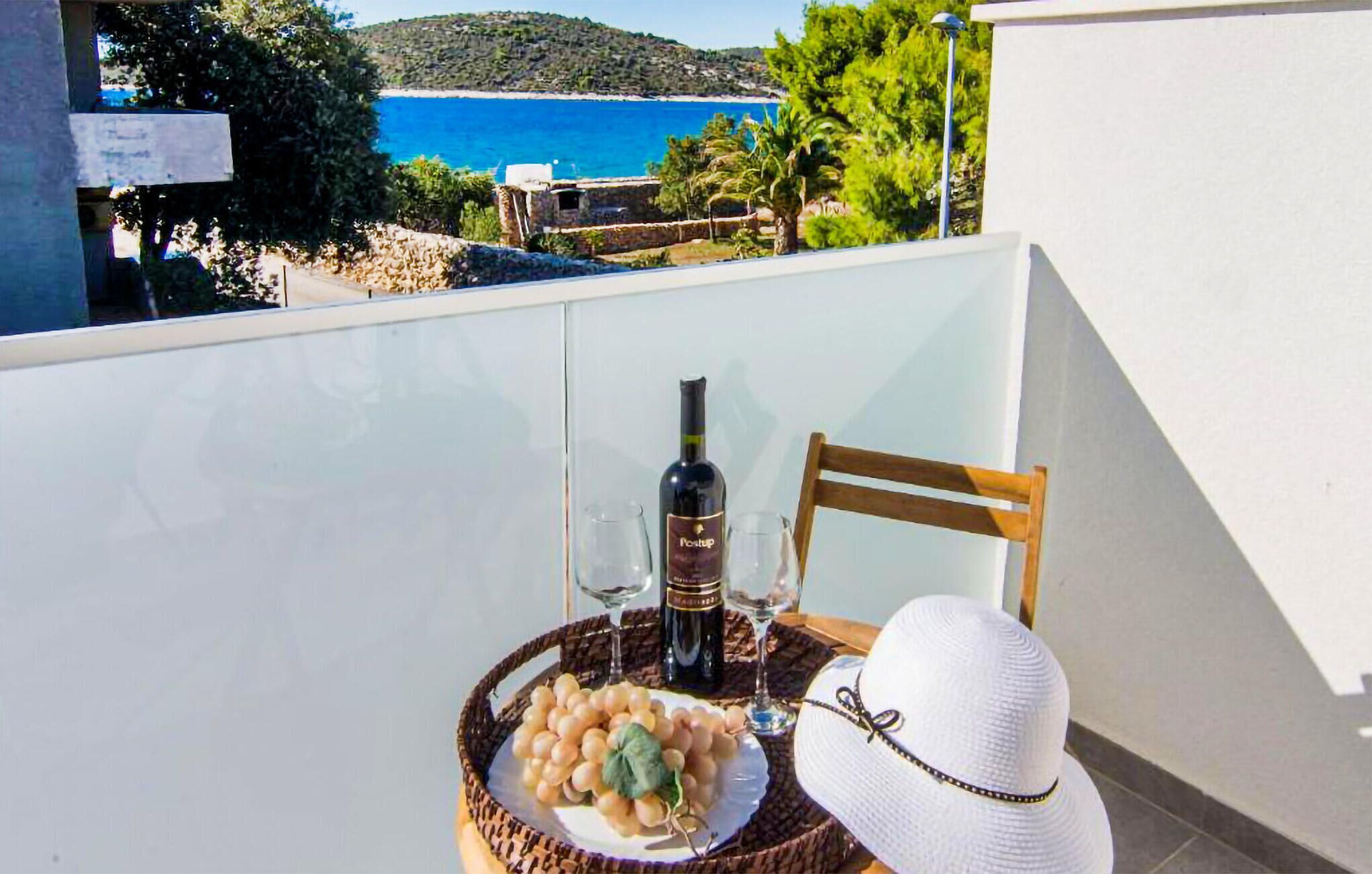Un balcone con un tavolo apparecchiato per due persone, con una bottiglia di vino, bicchieri, un cesto di uva e un cappello bianco.
